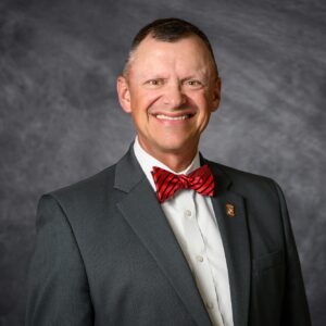 A man smiling wearing a suit and red bowtie against a gray background.