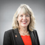 Lori Reesor is pictured indoors wearing a red dress and black jacket over it. Her hair is blond to her shoulders.