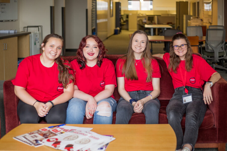 Four women sitting on a couch in an office smiling in red shirts promoting Badger Recovery.