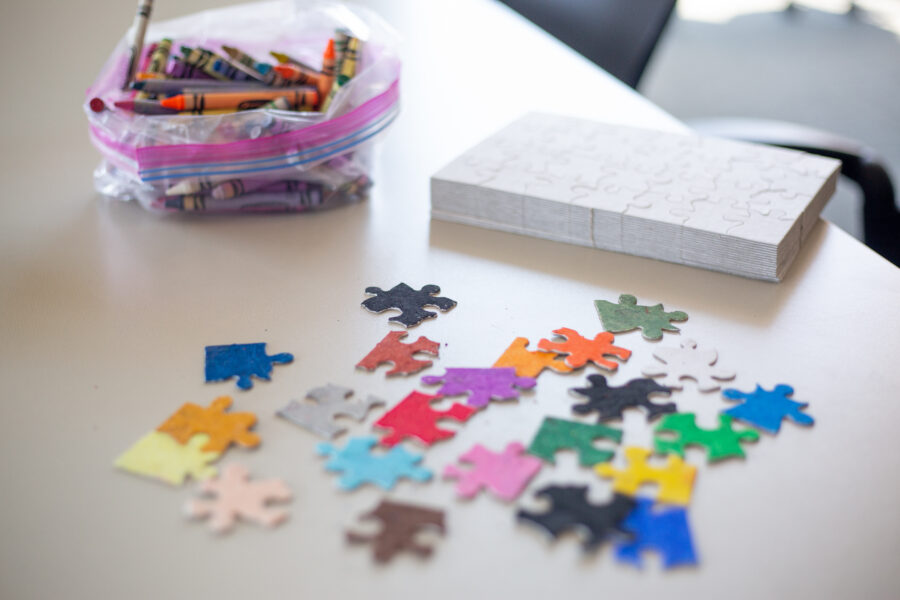 Multicolor puzzle pieces scattered on a table. There is a bag of crayons and white puzzles in the background.