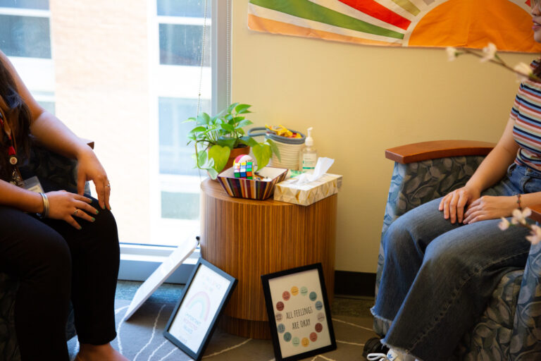 A student and a mental health provider sitting in conversation in a UHS office. There is a table with fidget toys, tissues, and a plant between them.