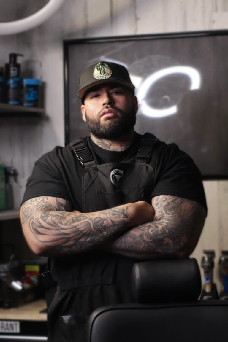 A man with a Bucks ball cap stands with his arms crossed behind a barbers chair.