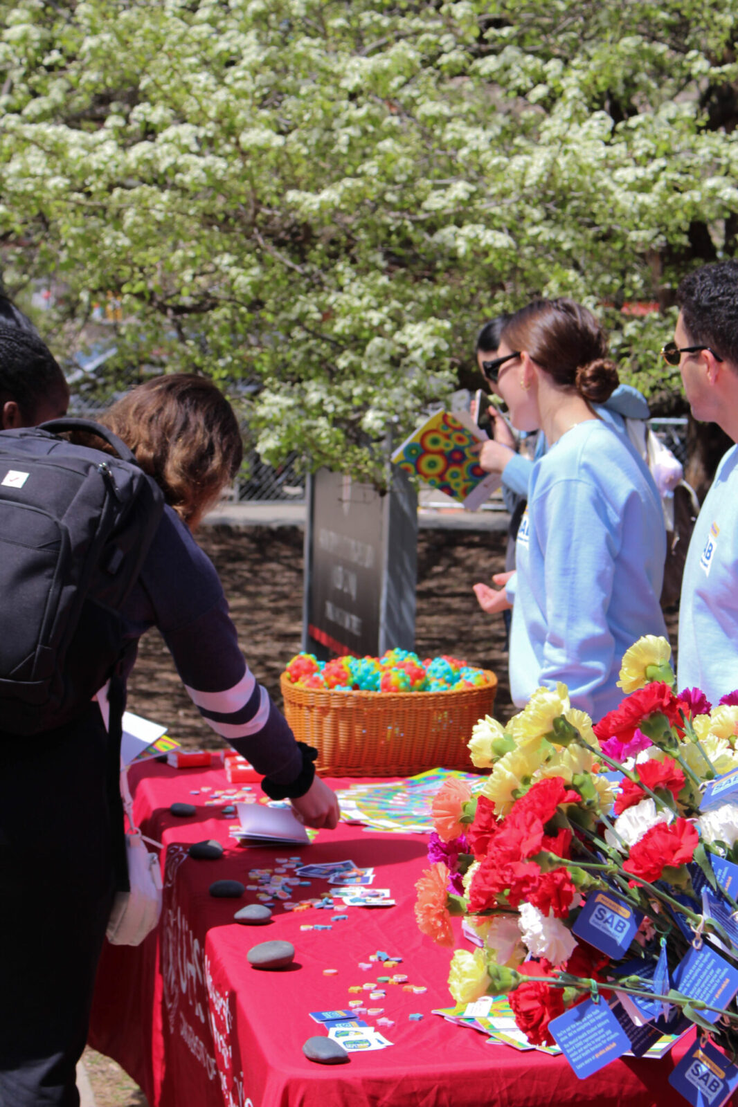 Student with black backpack and green cap grabbing an item from a basket at a tabling event. The table has markers postcards, and flowers.