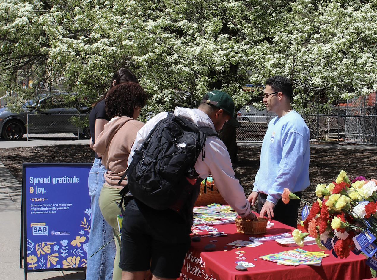 Student with black backpack and green cap grabbing an item from a basket at a tabling event. The table has markers postcards, and flowers.
