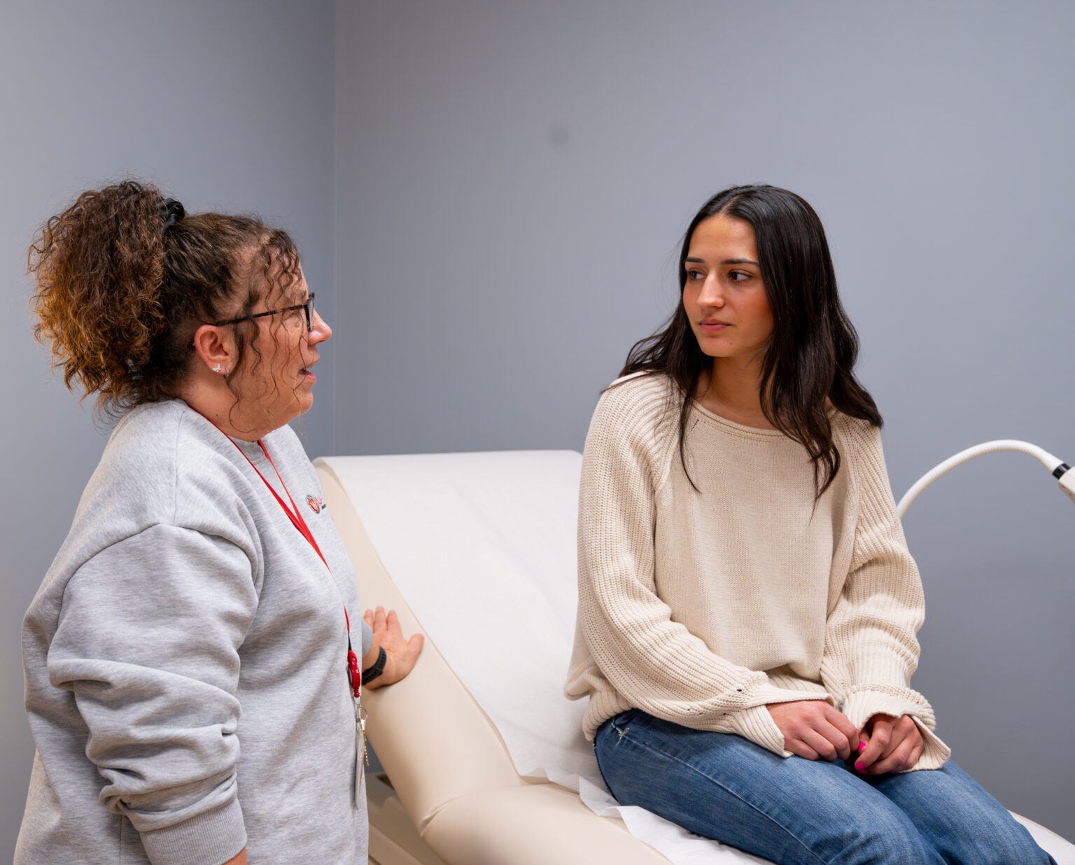A student sits on a medical table while listening to a medical provider.