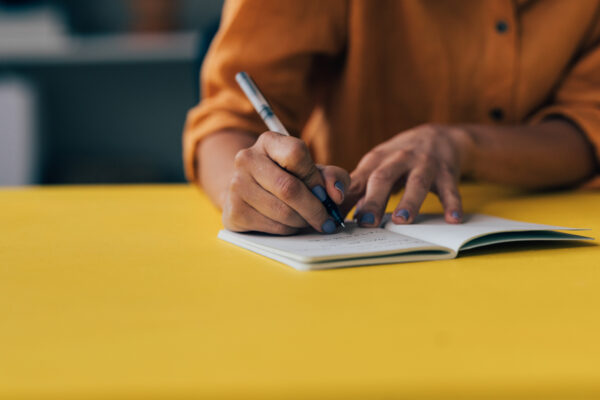 Focused shot of an individual's hands as they write notes in a notebook lying on a vibrant yellow surface.